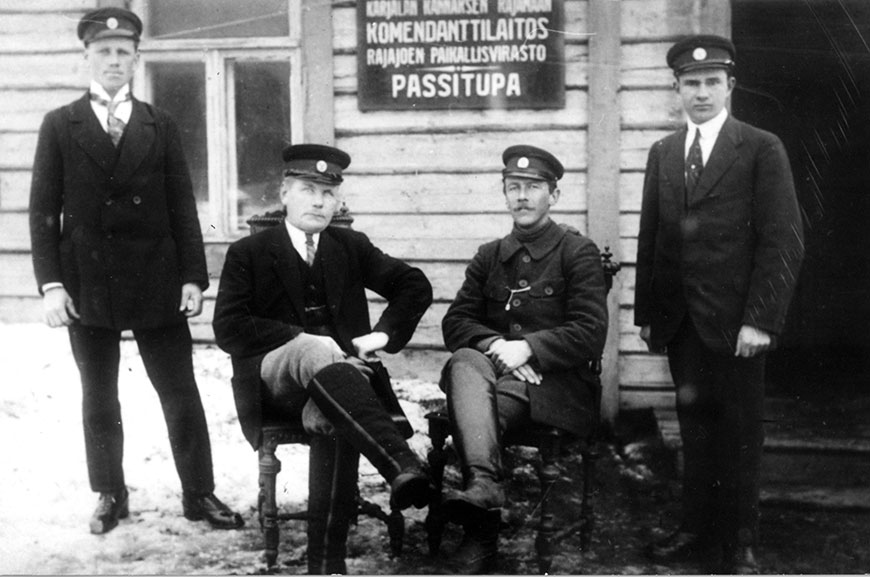 Neljä tullimiestä joista kaksi istuu ja kaksi seisoo Karjalan kannaksen Rajamaan komendanttilaitoksen Rajajoen paikallisviraston passituvan edustalla.  