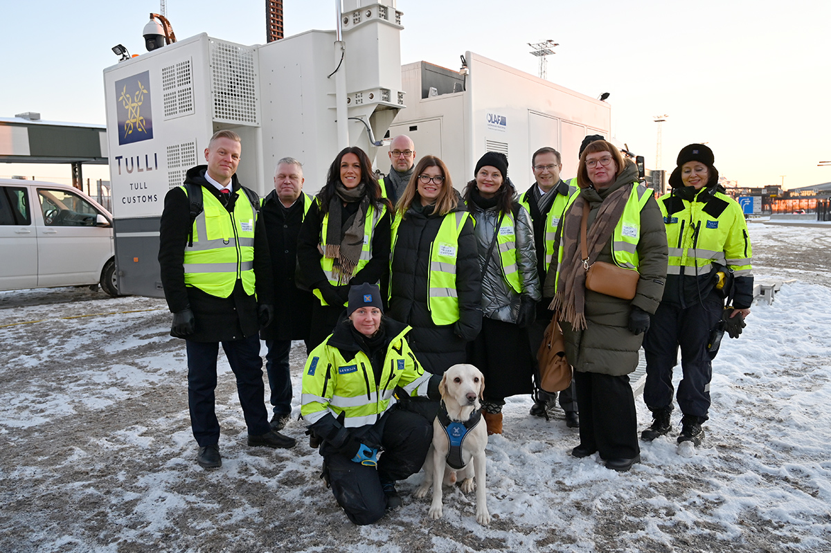 Ryhmäkuva hymyilevistä ihmisistä, Tullin logo taustalla, tullikoira edessä.