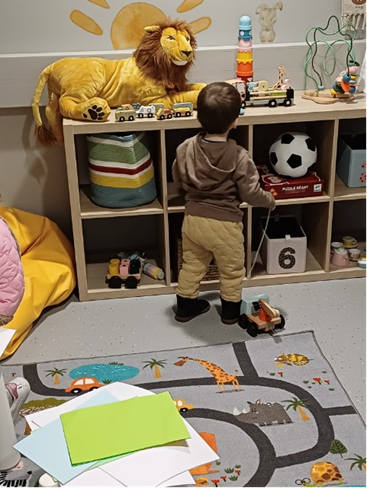 Pieni lapsi ruskeansävyisissä vaatteissa seisoo selin kameraan ja katsoo hyllyä, jossa on leluja. Lattialla on eläinkuosinen matto ja sen päällä piirustuspaperia.