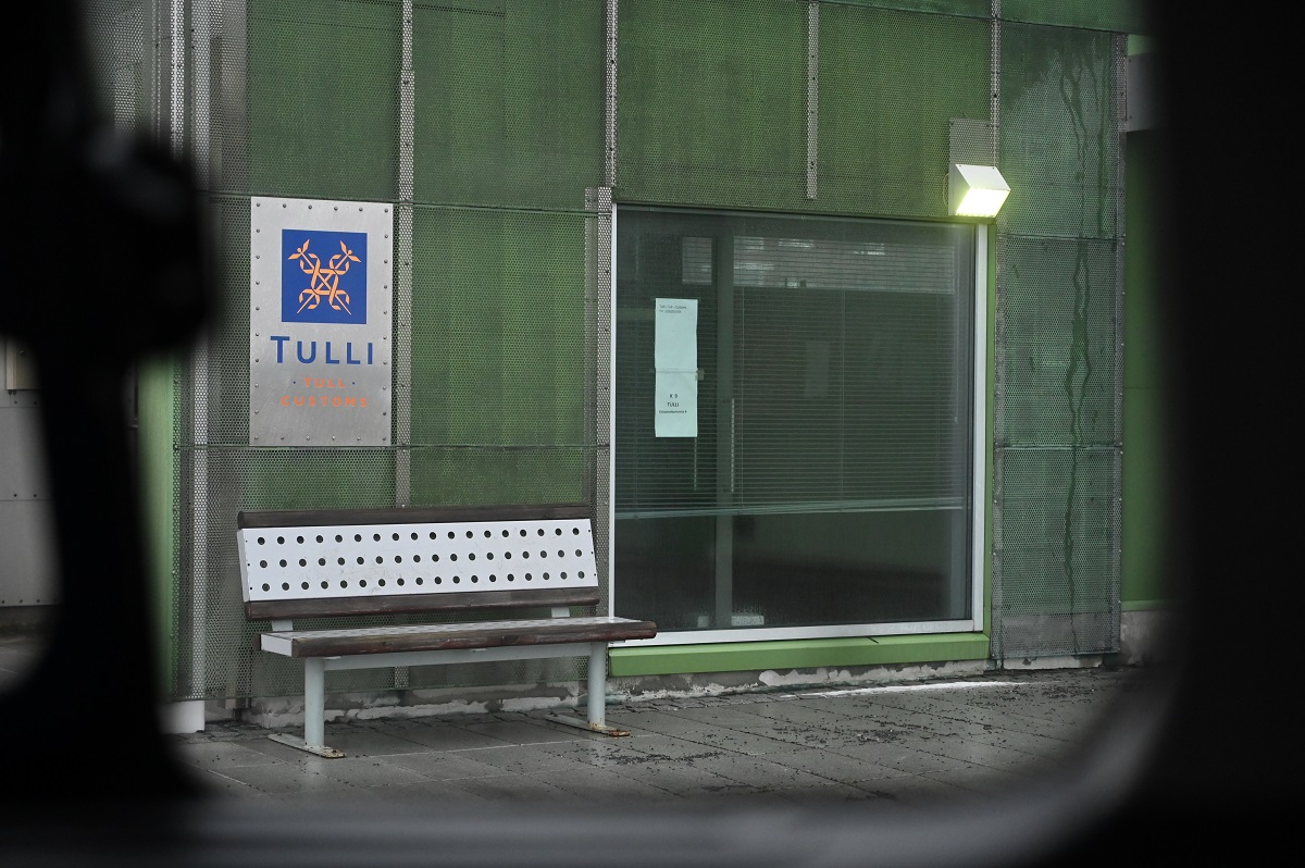 Kuva auton ikkunasta, josta näkyy vihreä seinä ja ikkuna, tyhjä penkki ja Tullin kyltti.