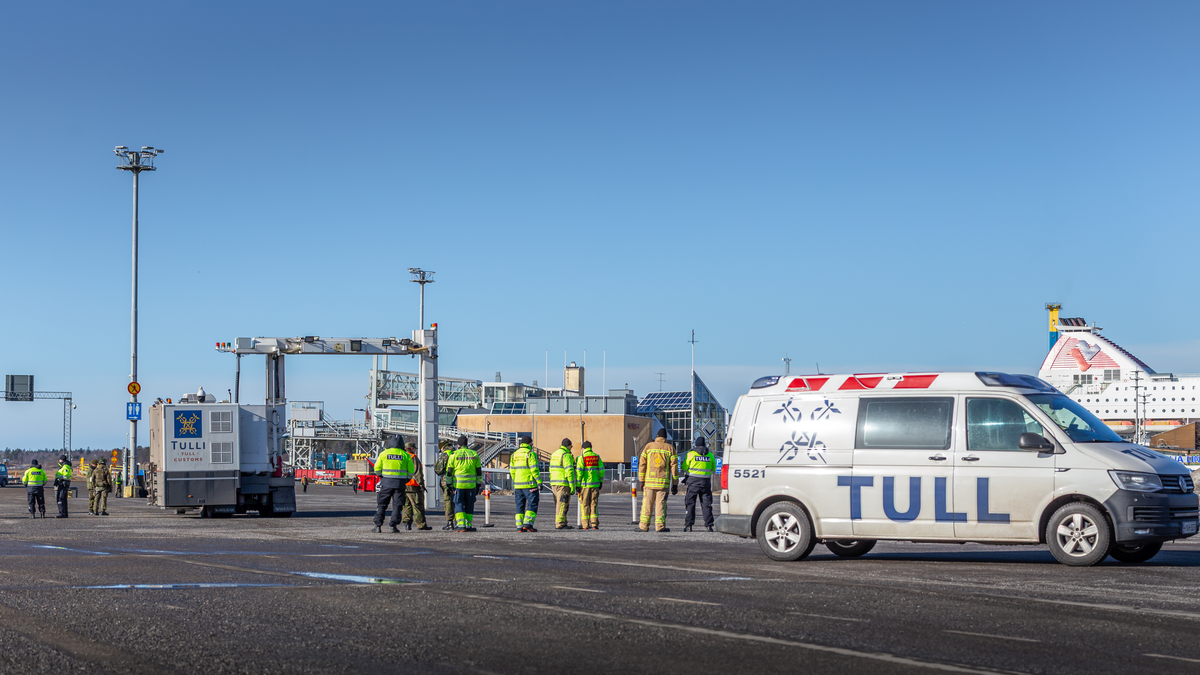 Tullin ajoneuvo, läpivalaisulaite ja henkilöstöä odottavat satamassa ajoneuvojen saapumista laivalta.
