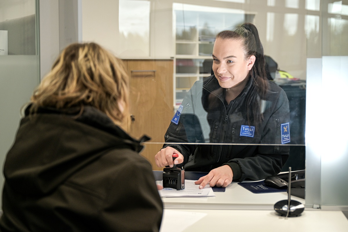 Tullin asiakaspalvelutiskillä tullivirkailija osoittaa paperista kohtaa asiakkaalle hymyillen leimasin kädessään.