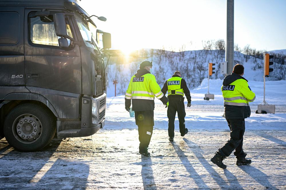 Kolme tullitarkastajaa aurinkoisessa ja lumisessa maisemassa kuorma-auton vierellä.