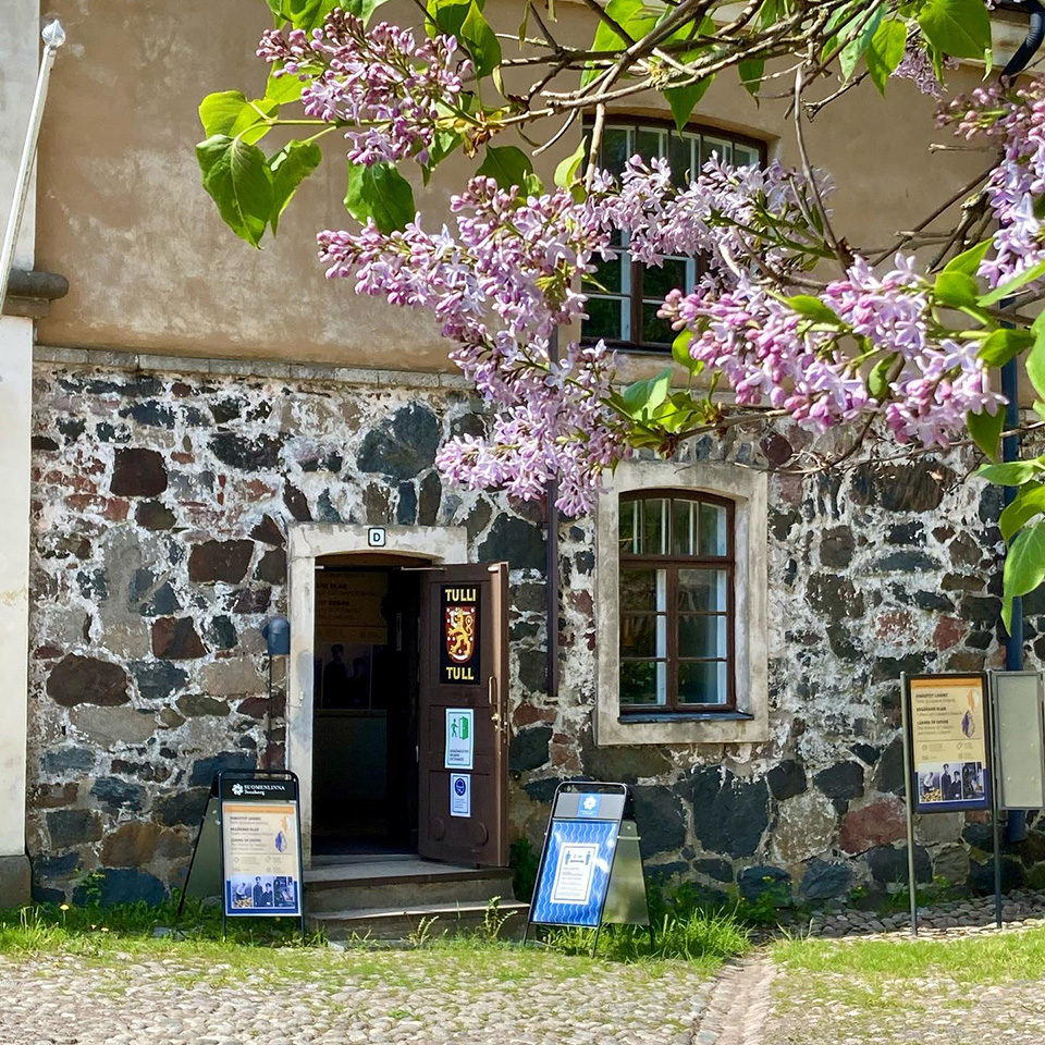 Etualalla syreenipuun oksia ja violetteja kukkia. Taustalla näkyy Tullimuseon kivinen rakennus, jonka ovi on auki. Pihassa on mukulakiveä.