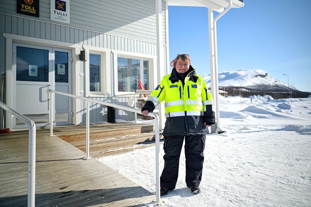 Tullivartija Marjut Muotkajärvi katsoo kameraan seisoen Kilpisjärven vaalean tullirakennuksen edessä lumisessa maisemassa.
