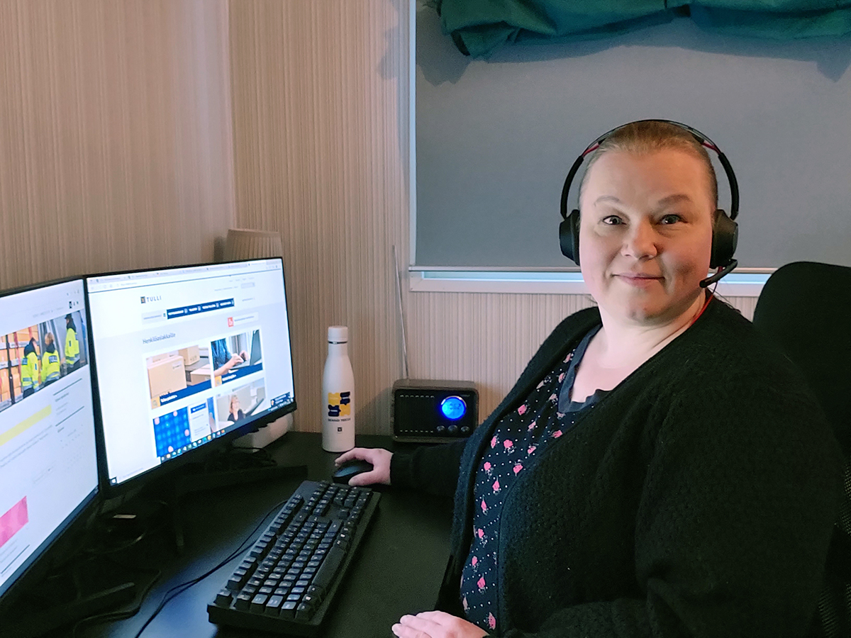 Nainen headset päässään istuu työpöydän ääressä kahden tietokoneruudun edessä ja katsoo kameraan.